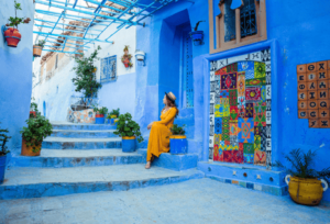 A scenic view of Chefchaouen’s blue-painted streets with a traveler in a vibrant yellow dress sitting on the steps, showcasing the charm of the 5 Days Tour from Casablanca.